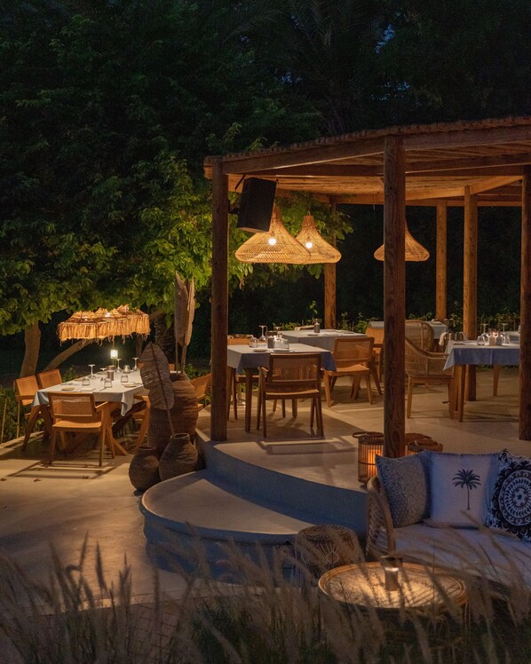 An outdoor dining area with wooden tables and chairs, hanging wicker lamps, and a covered seating area with cushions.