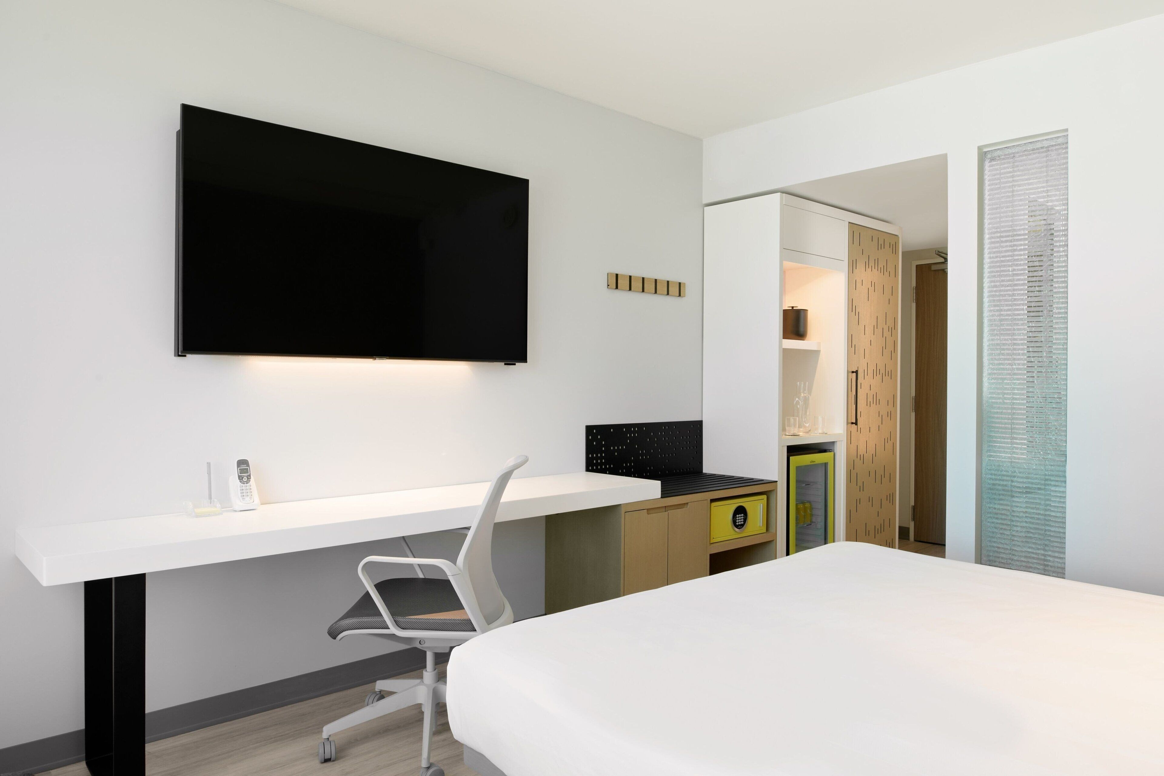A modern hotel room with a large bed, a desk with a chair, a flat-screen TV mounted on the wall, a wardrobe, and a bathroom with a shower and toilet.