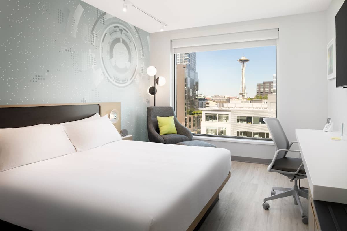 Habitación de hotel moderna con una cama grande, un escritorio y una silla. Se observa una vista de la ciudad a través de la ventana, incluyendo una torre.