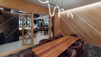 A modern conference room with a wooden table and chairs, a unique light fixture, and a view into another room with a staircase.