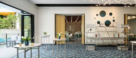 Un moderno vestíbulo de hotel con alfombra de diseño geométrico, mesas redondas con jarrones y una exhibición de vinos, con vistas al exterior.