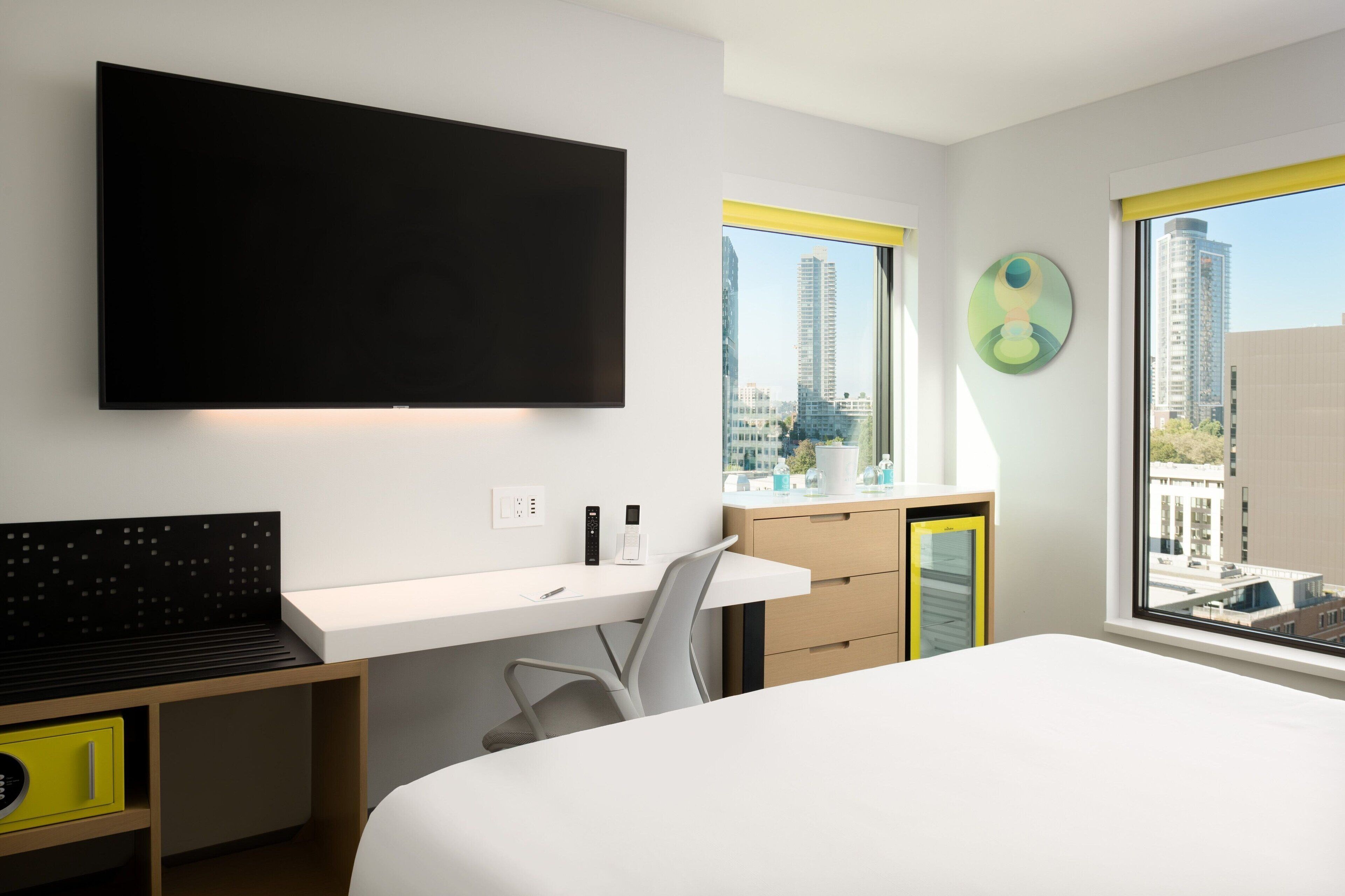 Une chambre d’hôtel moderne dotée d’un grand lit, d’un bureau avec une chaise, d’un téléviseur à écran plat fixé au mur et d’une fenêtre donnant sur la ville.