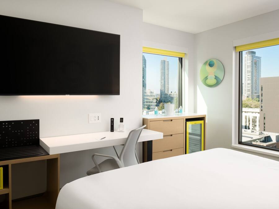 Une chambre d’hôtel moderne dotée d’un grand lit, d’un bureau avec une chaise, d’un téléviseur à écran plat fixé au mur et d’une fenêtre donnant sur la ville.