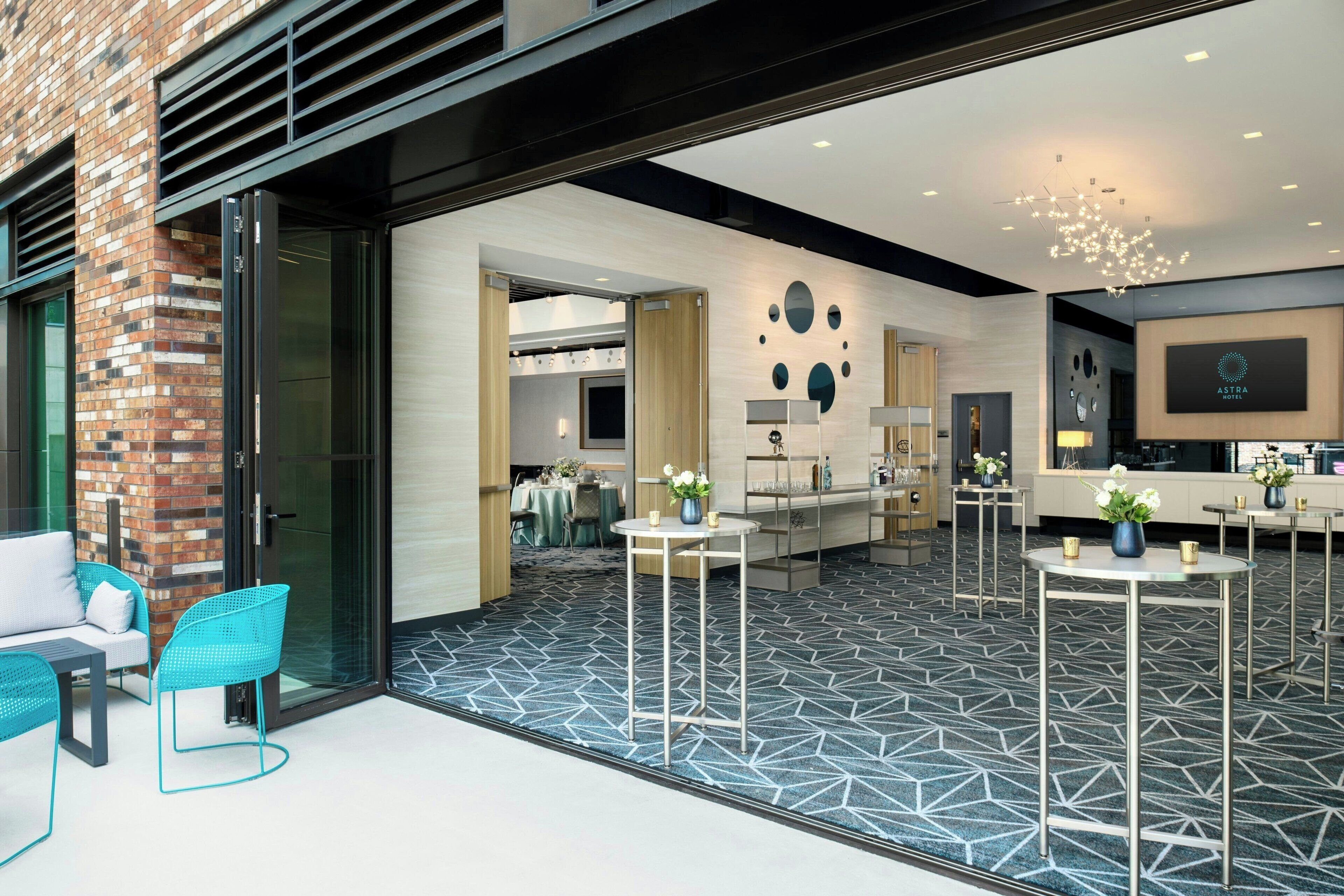 A modern hotel lobby with a geometric patterned floor, a seating area with blue chairs, and a reception desk with a chandelier.