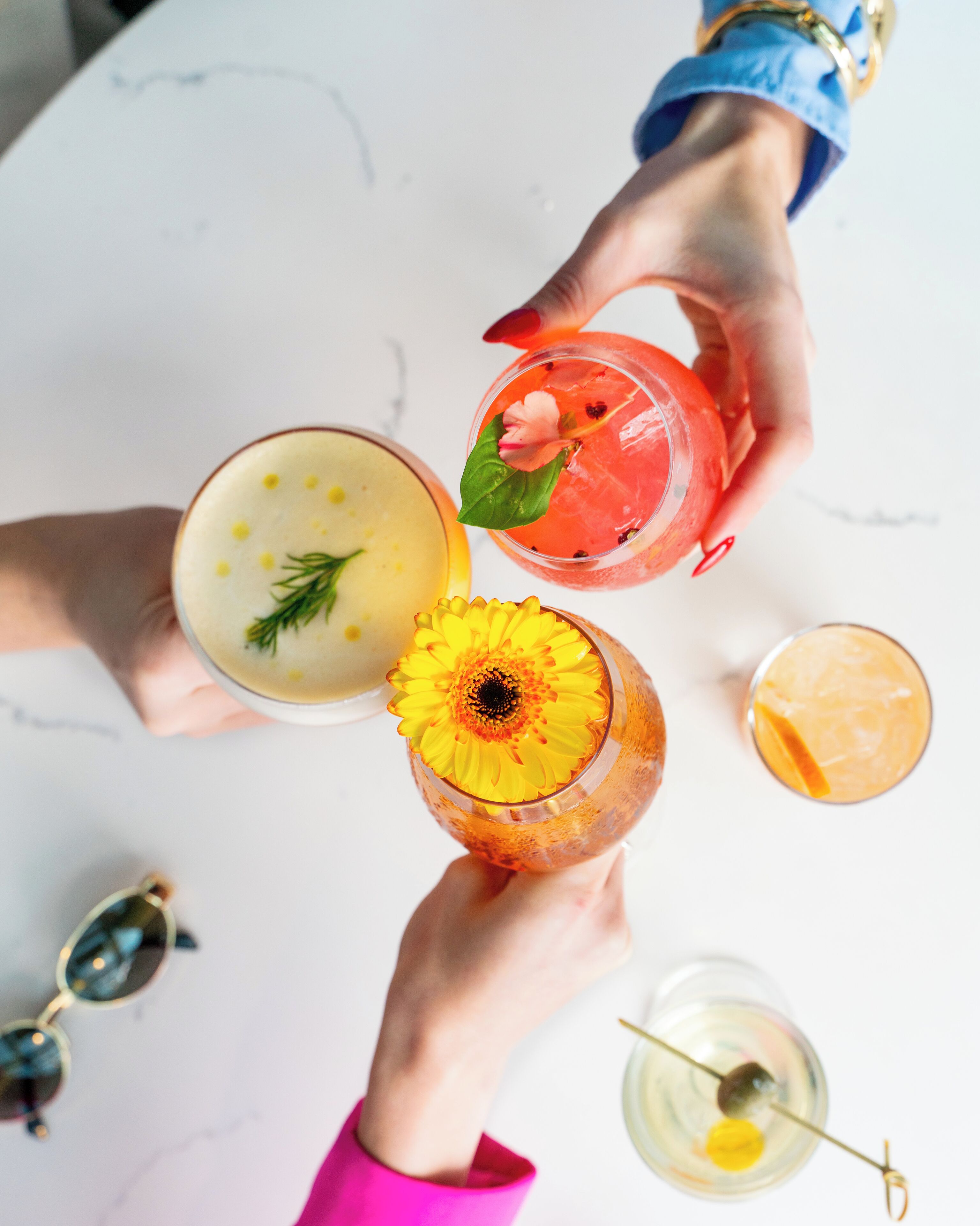 Von oben betrachtet, ein weißer Tisch mit verschiedenen Getränken, darunter eine Ananas-Garnitur und eine Blume, sowie ein Paar Sonnenbrillen.