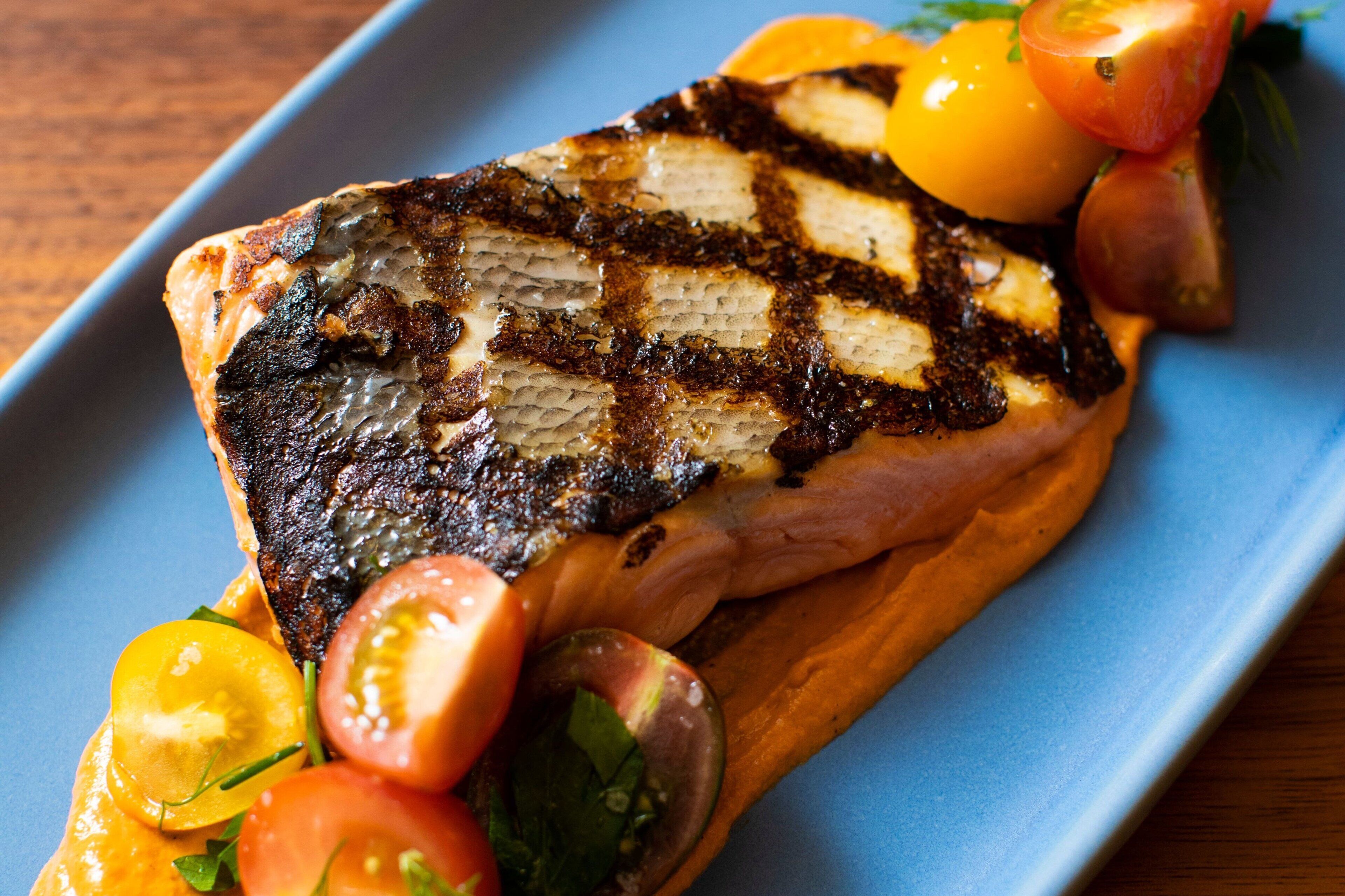A grilled salmon fillet with a charred pattern, accompanied by cherry tomatoes and a garnish of herbs, served on a blue plate.