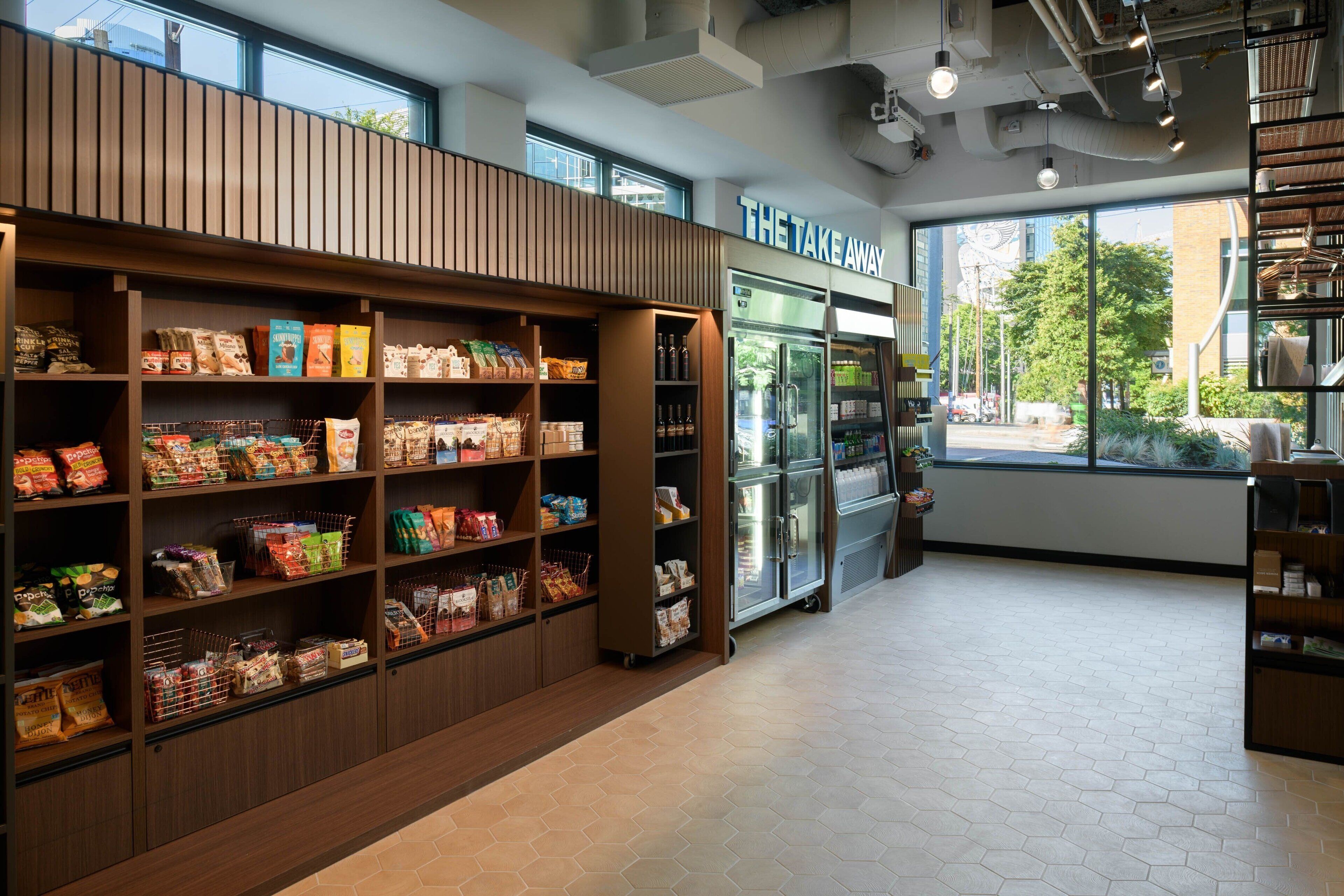 A grocery store interior with wooden shelving stocked with various food items, a refrigerated display case, and a large window providing a view of the outside.