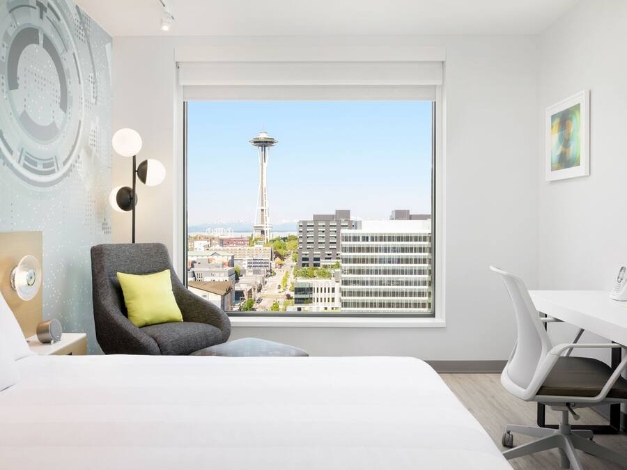 A hotel room with a bed, a desk, a chair, a lamp, and a view of the Seattle Space Needle.