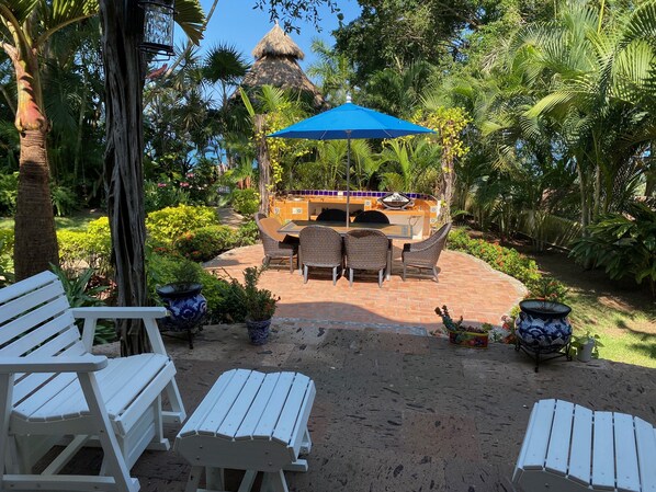 Un patio al aire libre con zona de barbacoa, rodeado de abundante vegetación y mobiliario exterior blanco.