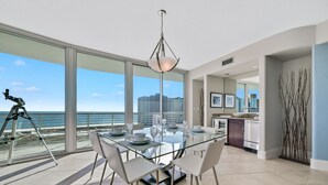 A modern dining area with a glass table and chairs, a city view, and a balcony with a tripod. - Bella Luna 810-Spectacular Views of Beach & Bay (Orange)