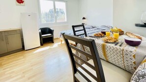 A small, bright room with a bed, a dining table with breakfast, a chair, a small kitchen area, and a window.