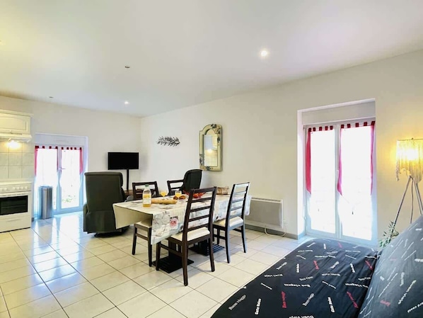 A dining area with a table set for four, a kitchen in the background, and a large window with curtains.