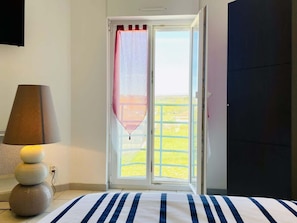 A room with a bed, a lamp, a window with a view of a balcony, and a door.