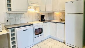 A compact kitchen with white cabinets, a built-in oven, a dishwasher, and a refrigerator. - Bonaparte (Bains-les-Bains)