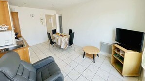 A living room with a sofa, a dining table with chairs, a television on a stand, and a kitchen area with a refrigerator and a microwave. - Victor Hugo (Bains-les-Bains)