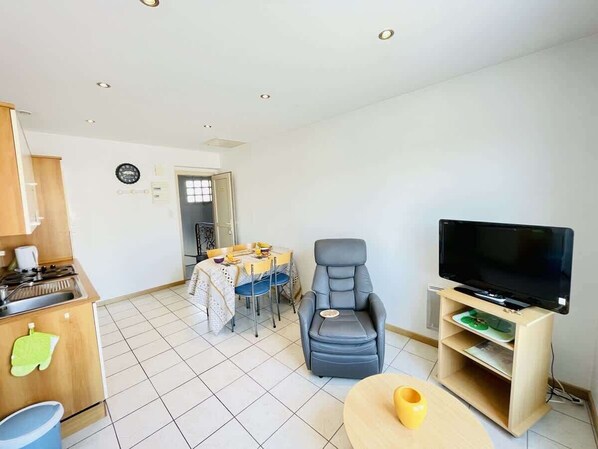 Un espace de vie compact comprenant un coin cuisine, un coin repas, un coin salon avec un téléviseur et une petite table.