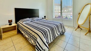 A bedroom with a bed, a nightstand, a mirror, and a window with a view. - Victor Hugo (Bains-les-Bains)