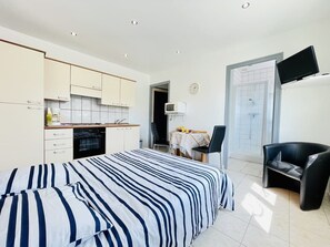 A bedroom with a bed, a desk, a chair, a TV, and a kitchenette.