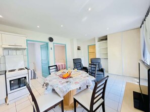 A dining area with a table, chairs, a clock, and a kitchen with a stove and cabinets. - Pasteur (Bains-les-Bains)