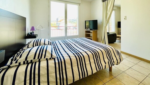 A hotel room with a bed, a television, a desk with a lamp, and a balcony with curtains.
