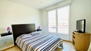 A bedroom with a bed, a TV on a wooden stand, a window with a view, and a nightstand with a lamp.