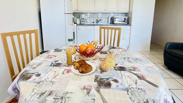 A dining area with a table set for two, a refrigerator, and a kitchen in the background. - Porthos (Bains-les-Bains)