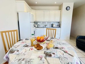 A dining area with a table set for two, a refrigerator, and a kitchen in the background. - Porthos (Bains-les-Bains)