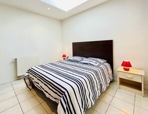 A bedroom with a bed, a nightstand, a lamp, a radiator, and a skylight. - Porthos (Bains-les-Bains)