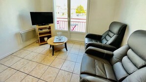 A living room with a TV, a sofa, a small table, and a balcony with a railing. - Jacques Prévert (Bains-les-Bains)