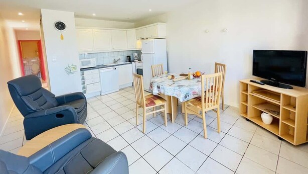 Un salon avec un canapé, une table à manger entourée de chaises, un téléviseur posé sur un meuble en bois, et une cuisine à l’arrière-plan.