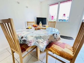 A dining area with a round table, four wooden chairs, a television, and a window with curtains. - Aramis (Bains-les-Bains)