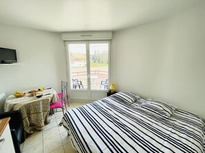 Une chambre avec un lit, un bureau, une chaise, une table avec le petit-déjeuner, et un balcon avec des chaises.