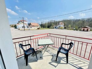 A balcony with two chairs and a table, surrounded by a red railing, overlooking a parking lot and buildings.