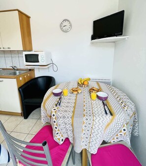 A compact kitchen with a microwave, a dining table with a patterned tablecloth, a clock, and a flat-screen TV on the wall.