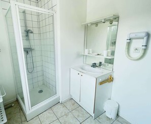 A bathroom with a glass-enclosed shower, a white sink with a mirror above it, and a white trash bin. - Jean de la Fontaine : Studio proche des Thermes (Bains-les-Bains)