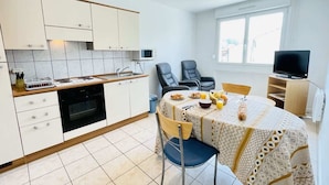 Une cuisine moderne avec des meubles blancs, une table à manger recouverte d’une nappe à motifs et un téléviseur placé dans un coin.