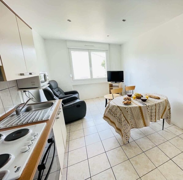 A modern kitchen with a stove, sink, and microwave, a dining area with a table set for two, and a TV in the background.