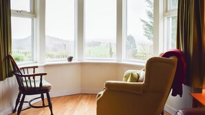 Ein Zimmer mit Erkerfenster, einem Holzstuhl, einer Couch und Blick auf eine grüne Landschaft.