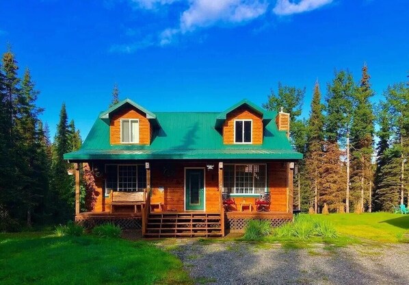 A two-story log cabin with a green roof, a porch with seating, and a small front yard. - Kenai River Getaway (Soldotna)