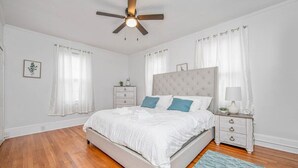 Un dormitorio con cama, mesita de noche, ventilador de techo y ventana con cortinas.