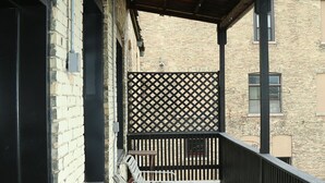 A covered outdoor area with a wooden floor, two white metal chairs, a black metal fence, and a brick building in the background.