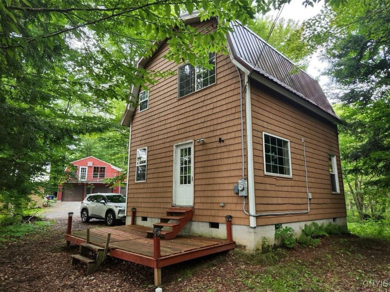 Une cabane en bois avec un porche à l’avant, un garage et un bâtiment rouge en arrière-plan.