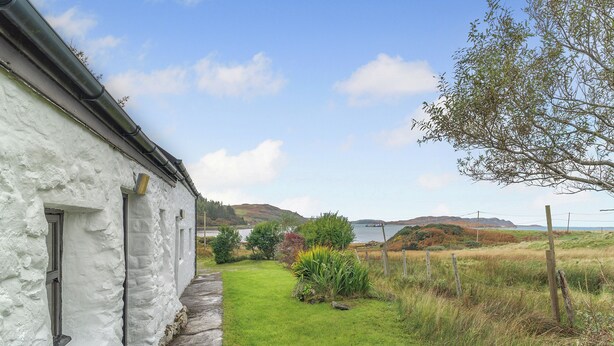 A white building with a narrow pathway leading to a grassy area with a view of the sea and hills in the distance. - A tiny, traditional cottage tucked away on the Ross of Mull, close to the sea. (Isle of Mull)