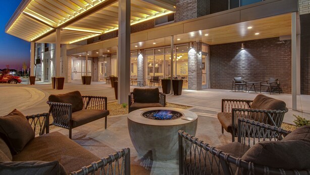 Un espace extérieur aménagé avec des chaises en osier et un foyer central, entouré d’une terrasse couverte dotée d’un mur en briques et de grandes fenêtres.