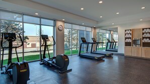 Une salle de sport équipée de tapis de course, de vélos elliptiques et d’un rameur, avec de grandes fenêtres et une horloge au mur.