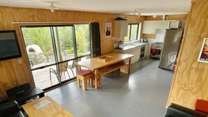 Amplia sala de estar con un sofá de cuero negro, una mesa de centro de madera, y un comedor con mesa y sillas de madera. Se ve una cocina con gabinetes blancos y un refrigerador al fondo.