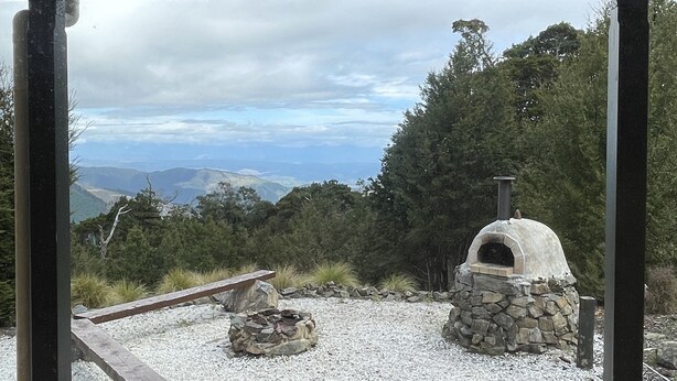 Un horno de piedra al aire libre con chimenea, rodeado de grava y rocas, con árboles y montañas lejanas de fondo.