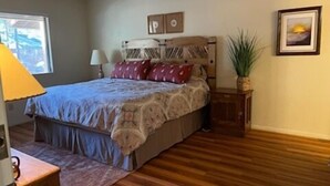 A bedroom with a bed, a ceiling fan, a nightstand, a lamp, a framed picture, and a potted plant.