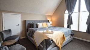 Ein Schlafzimmer mit einem Bett, einem Sessel, einem Nachttisch und einem Fenster mit Vorhängen.