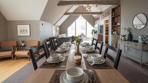 Un espace repas avec une longue table dressée pour six personnes, entourée de chaises, et offrant une vue sur l’extérieur grâce à de grandes fenêtres.
